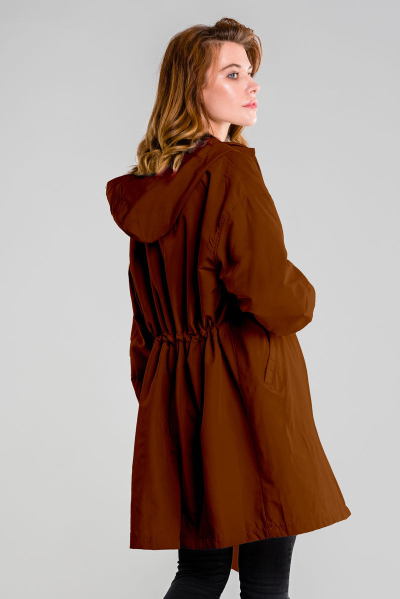 A person with long hair stands facing away, wearing the looselyboho BURNT BROWN WINDBREAKER. This dark brown hooded coat is crafted from lightweight, water-resistant fabric and features gathered detailing at the back. Its adjustable hood frames their side-turned gaze against a plain light gray background.