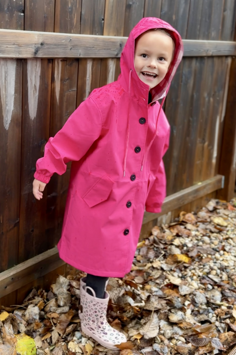 A child in the looselyboho Bright Berry Kids’ Windbreaker smiles outdoors on fallen leaves next to a wooden fence, pairing the water-resistant jacket with patterned rain boots.