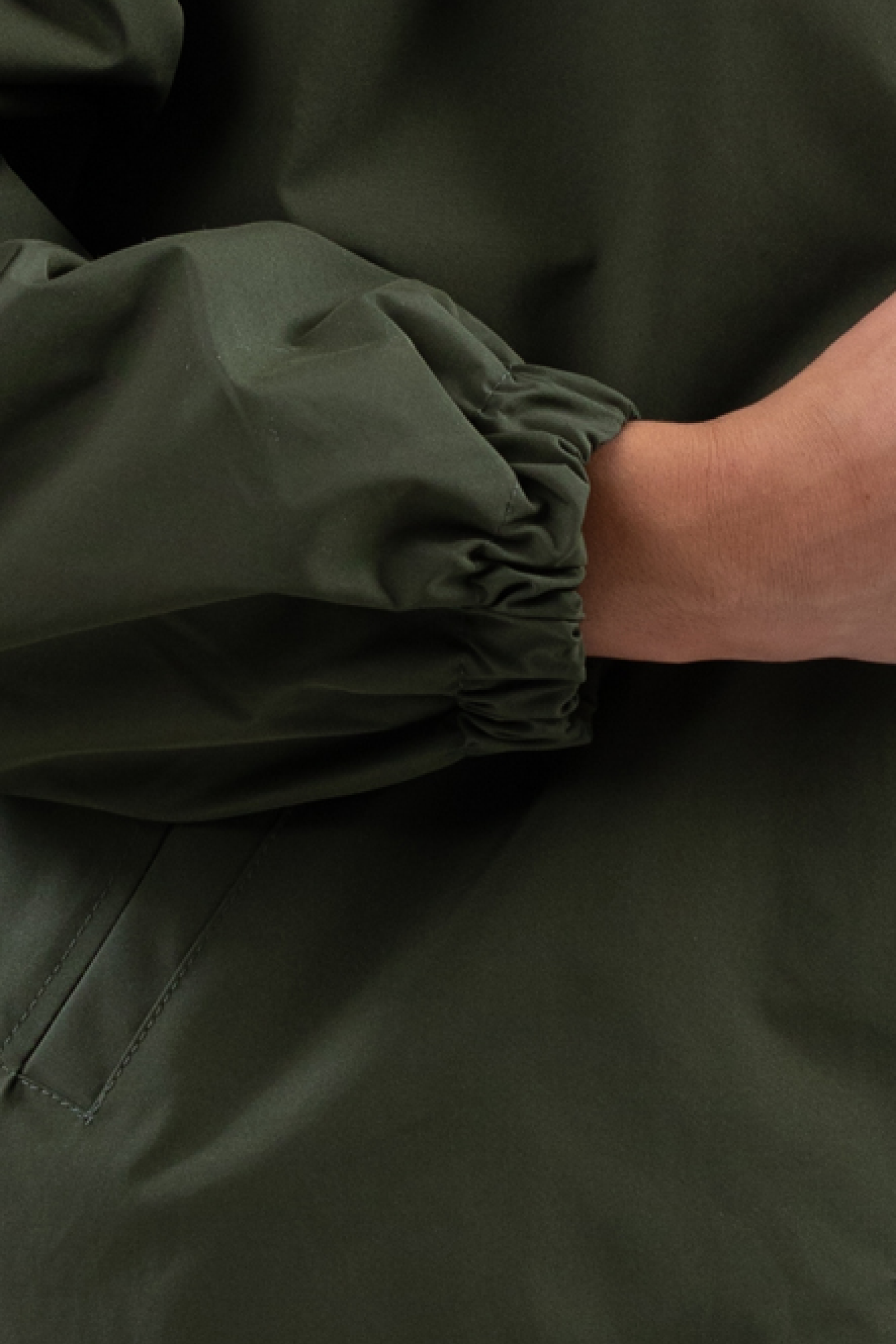 A close-up shows part of a person's arm and hand resting across their body, wearing the Icon Windbreaker Rain Jacket in olive green with elasticated cuffs.
