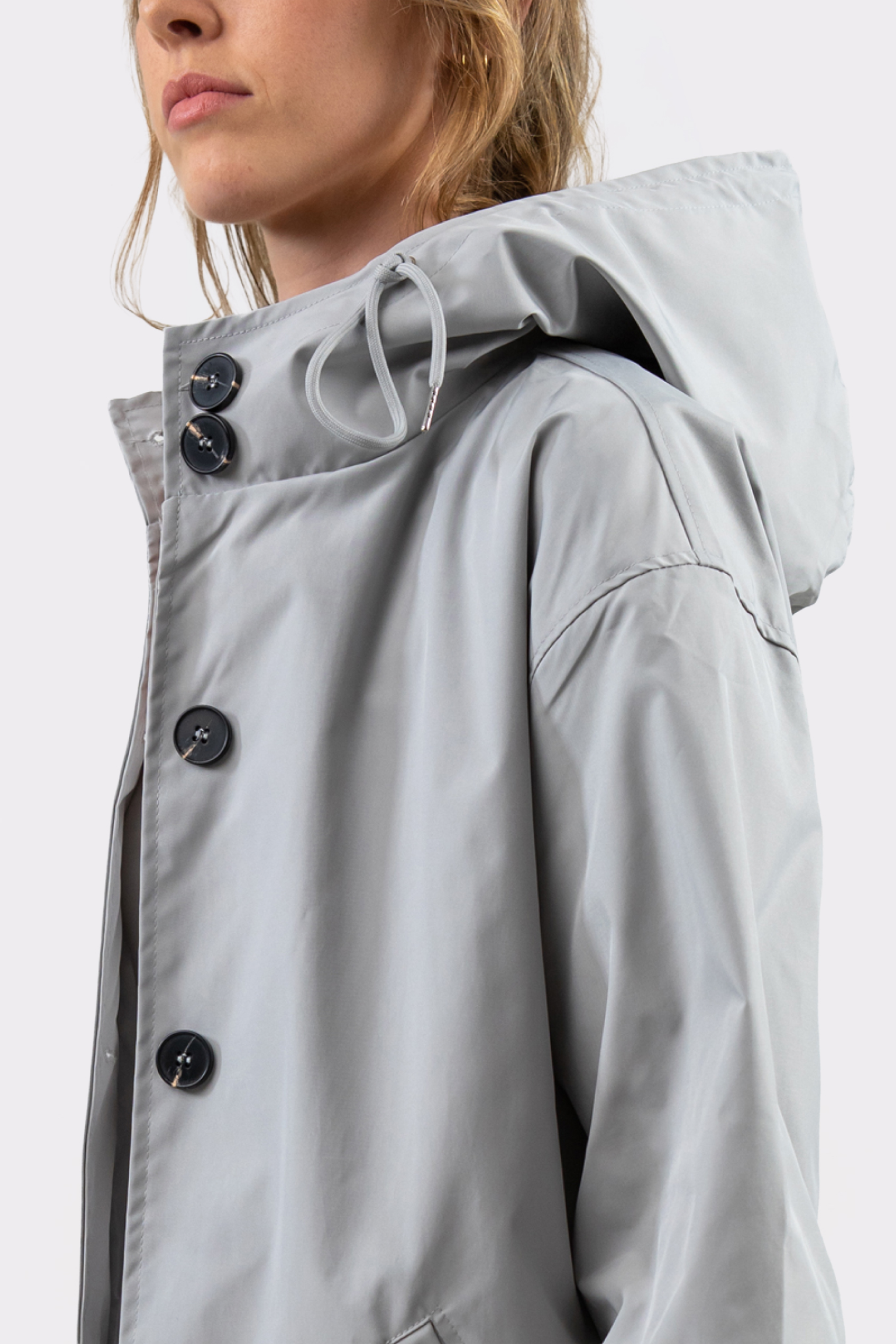 Someone in the Icon Windbreaker Rain Jacket—a light gray, matte-finish hooded raincoat with black buttons—shown from the shoulders up and facing sideways. The hood is up but not covering the head.