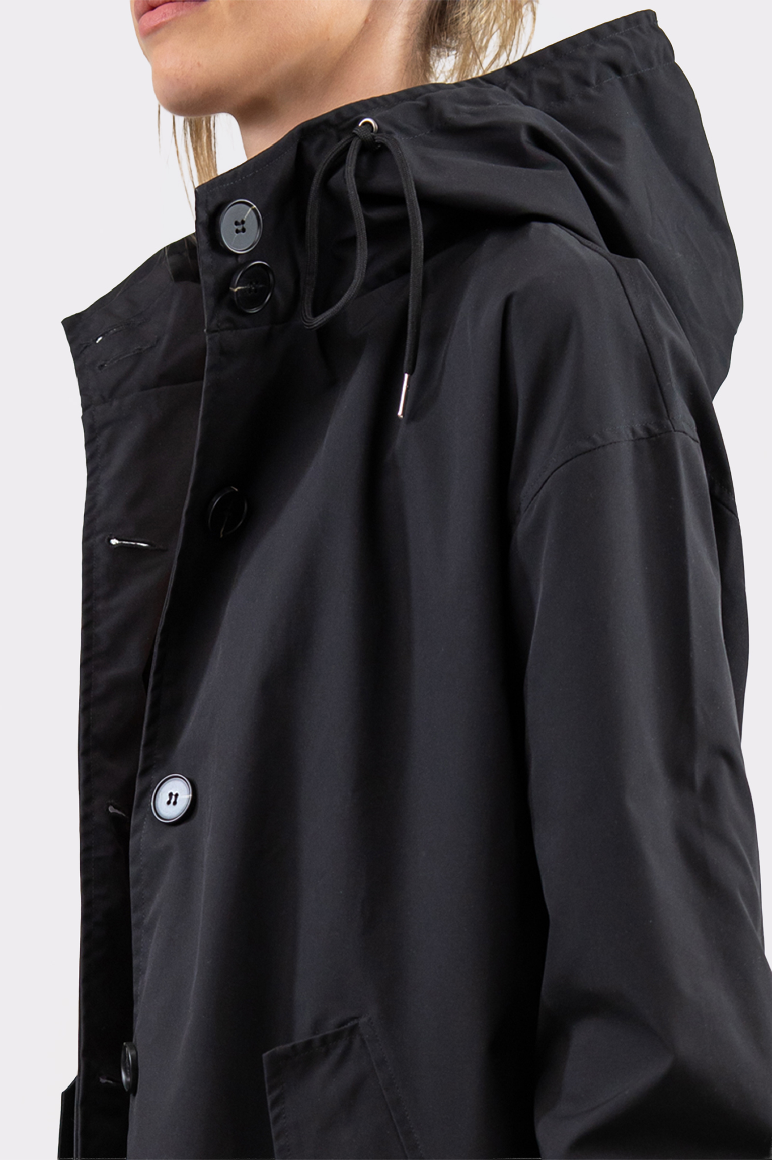 A person in a black Icon Windbreaker Rain Jacket with a hood, drawstring, large black buttons, and a flap covering the buttons, shown from the shoulders up in side profile.
