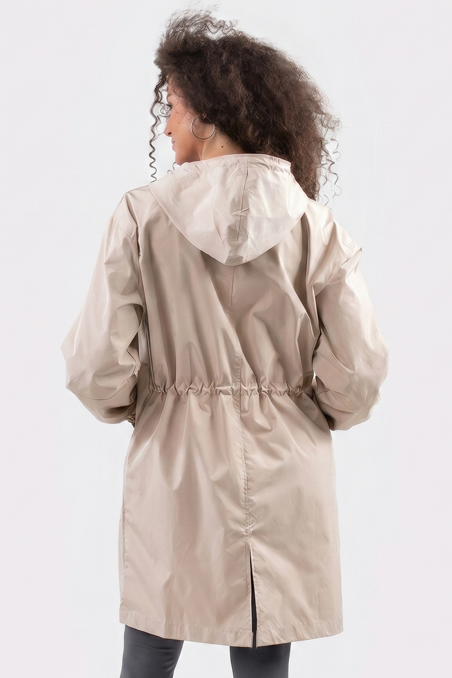 A person with curly hair wears the Icon Windbreaker Rain Jacket in light beige, shown from the back. The raincoat features a hood, drawstring waist, center seam, and a small bottom slit against a plain white background.