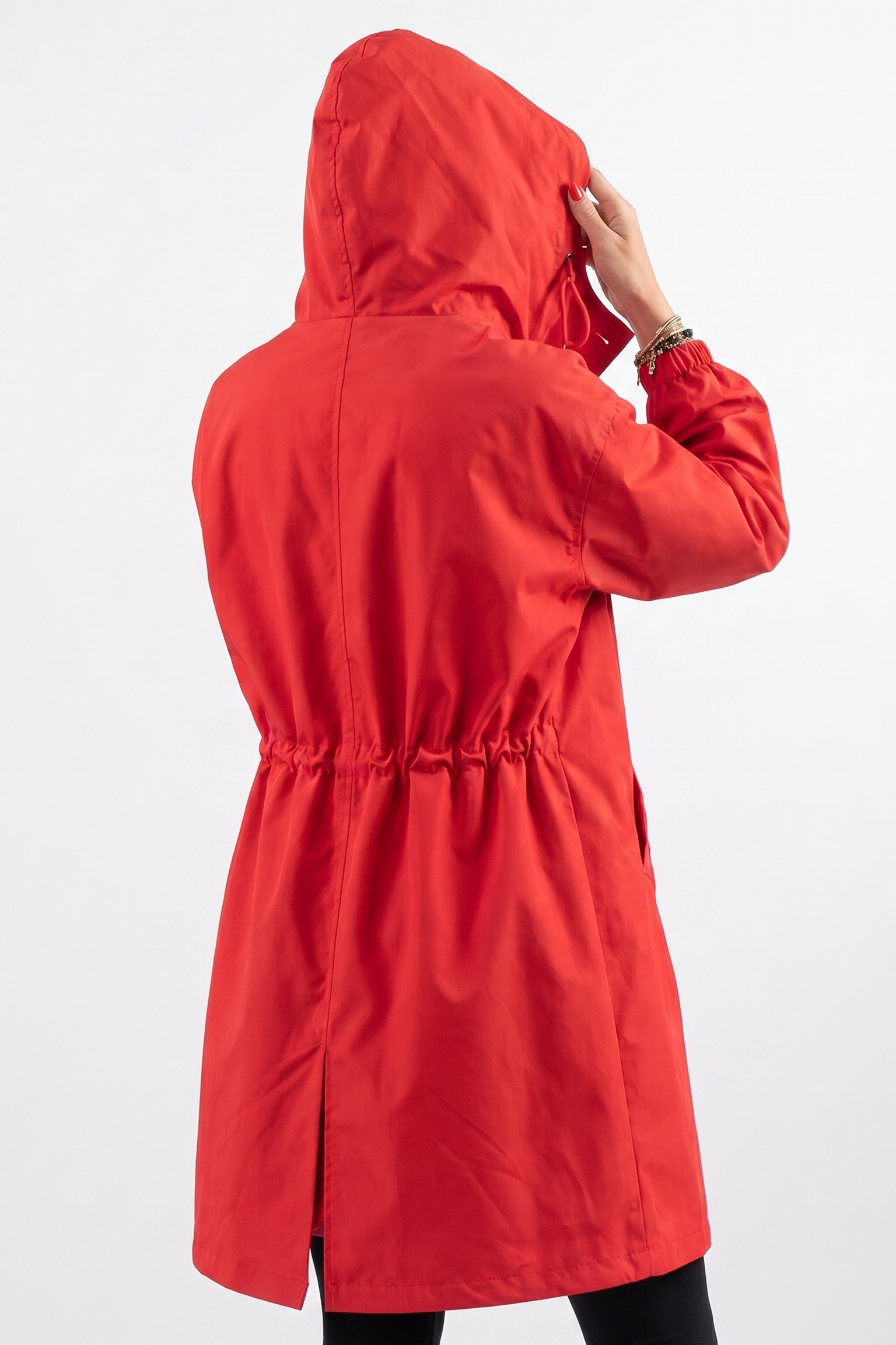 A person wearing the Icon Windbreaker Rain Jacket in bright red, featuring a hood, elastic waist, and back slit, stands facing away from the camera on a plain white background, adjusting the hood with one hand.
