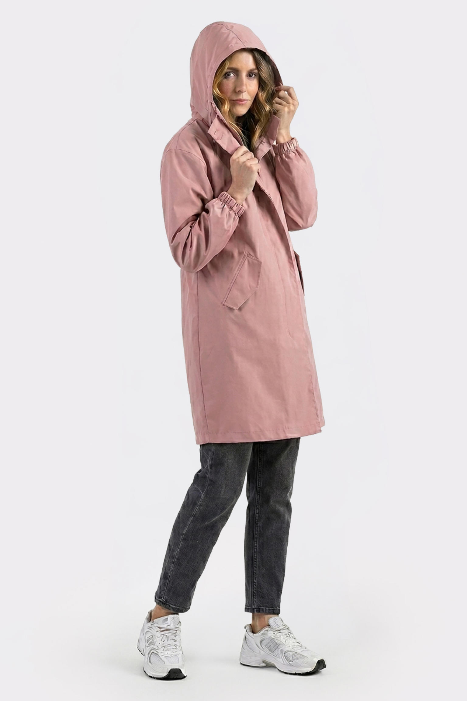 A person models the Icon Windbreaker Rain Jacket in pink, paired with black jeans and white sneakers, standing against a plain light background and touching the hood while facing the camera.