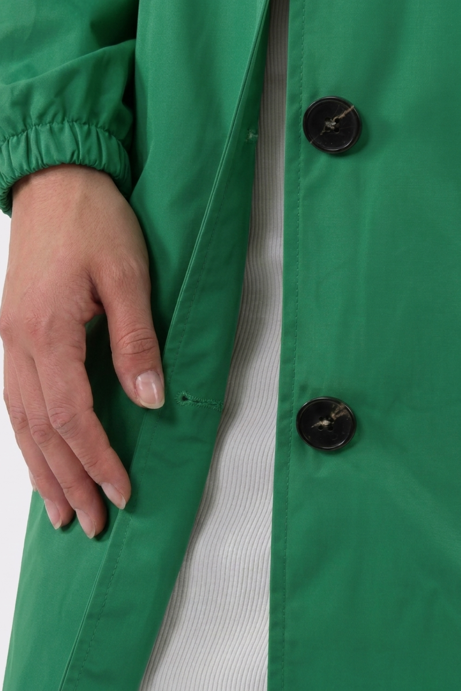 A close-up of a hand resting on the side of the green Icon Windbreaker Rain Jacket with large black buttons, worn over a white ribbed garment.