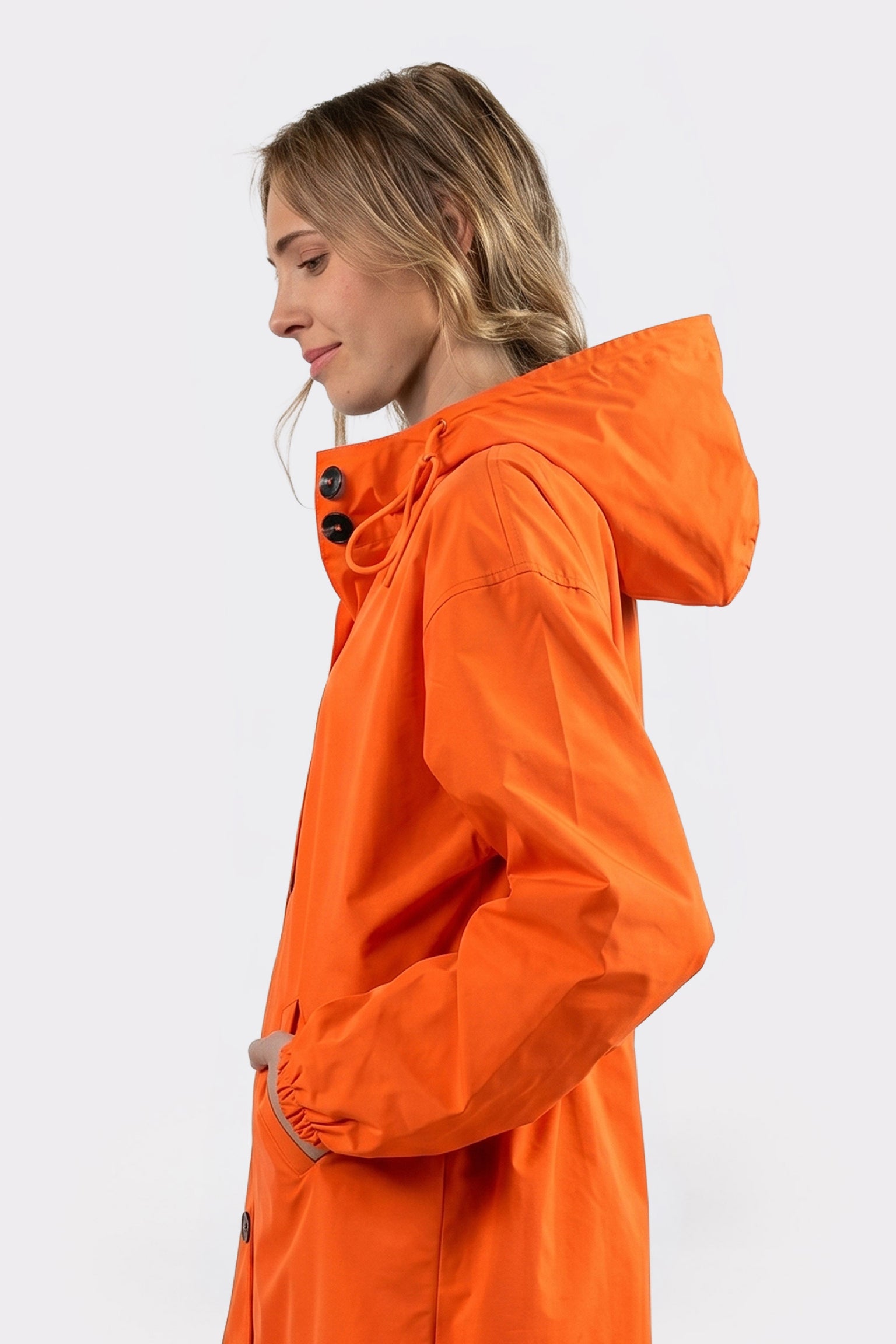 A person models the Icon Windbreaker Rain Jacket in orange, shown in profile with their left hand in their pocket and the hood down, against a plain light background.