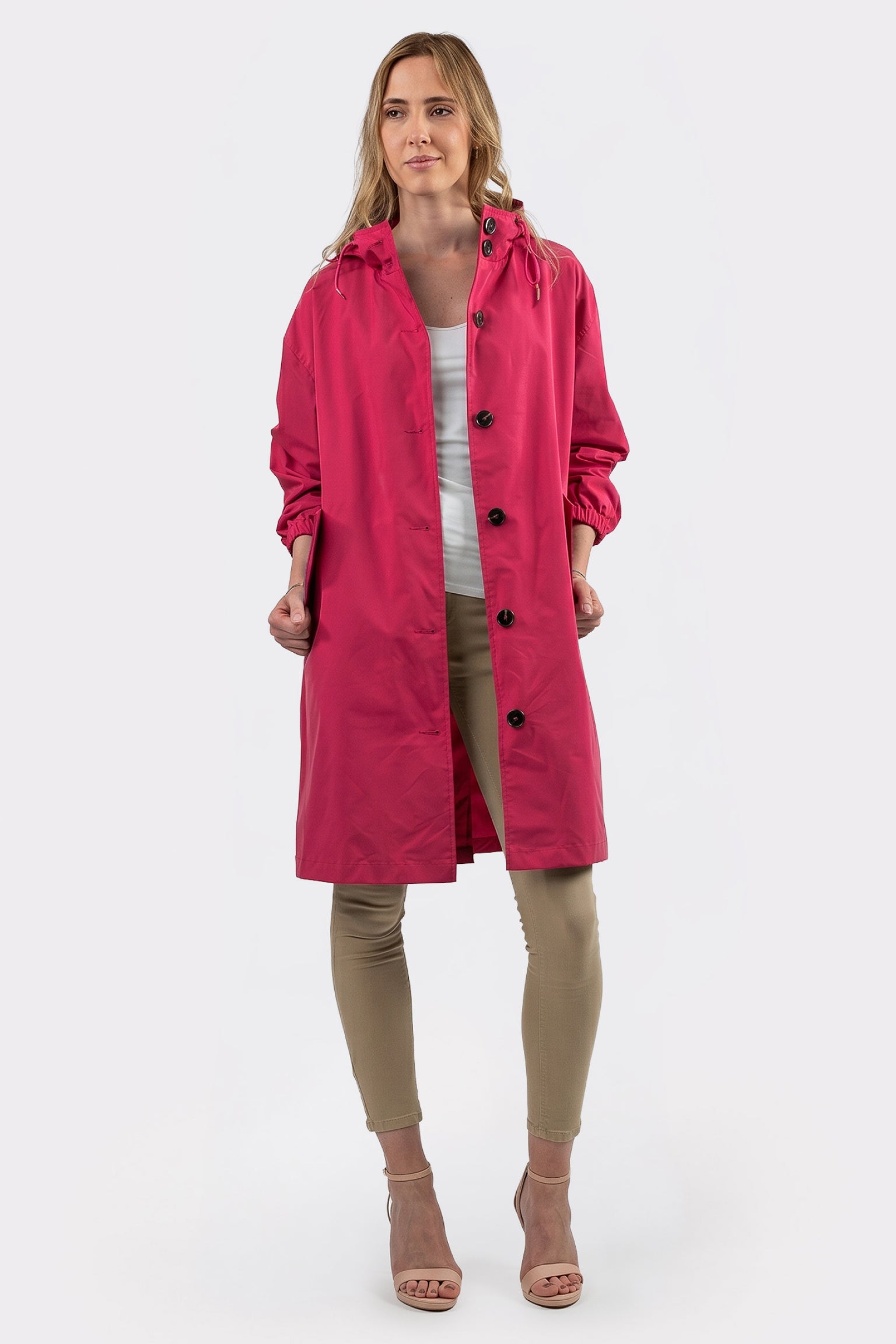 A woman faces forward in the Icon Windbreaker Rain Jacket, a bright pink knee-length coat, layered over a white top and beige pants with nude open-toed heels. She holds the sides of the unbuttoned jacket against a plain white background.