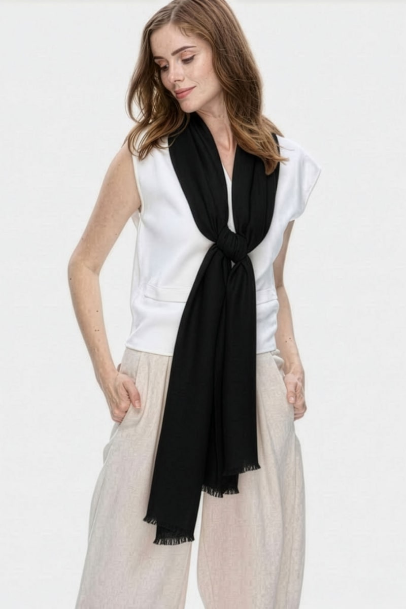 A person wears a sleeveless white top, light beige pants, and the looselyboho Noir Eclipse 100% Cashmere Scarf knotted loosely around their neck. Their hands are in their pockets, and the background is plain and light-colored.