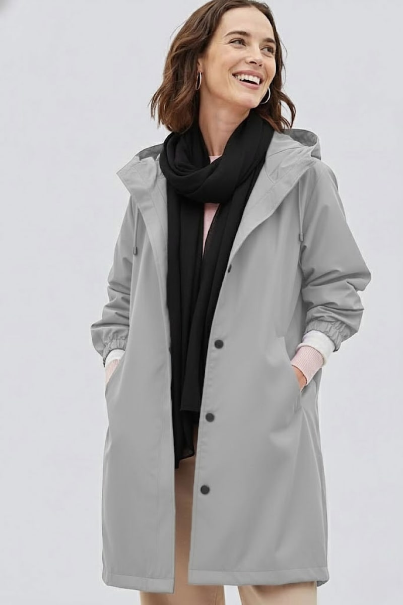 A woman wearing a light gray hooded raincoat, light-colored pants, and the looselyboho Noir Eclipse 100% Cashmere Scarf stands against a plain background, smiling and looking slightly to her left.