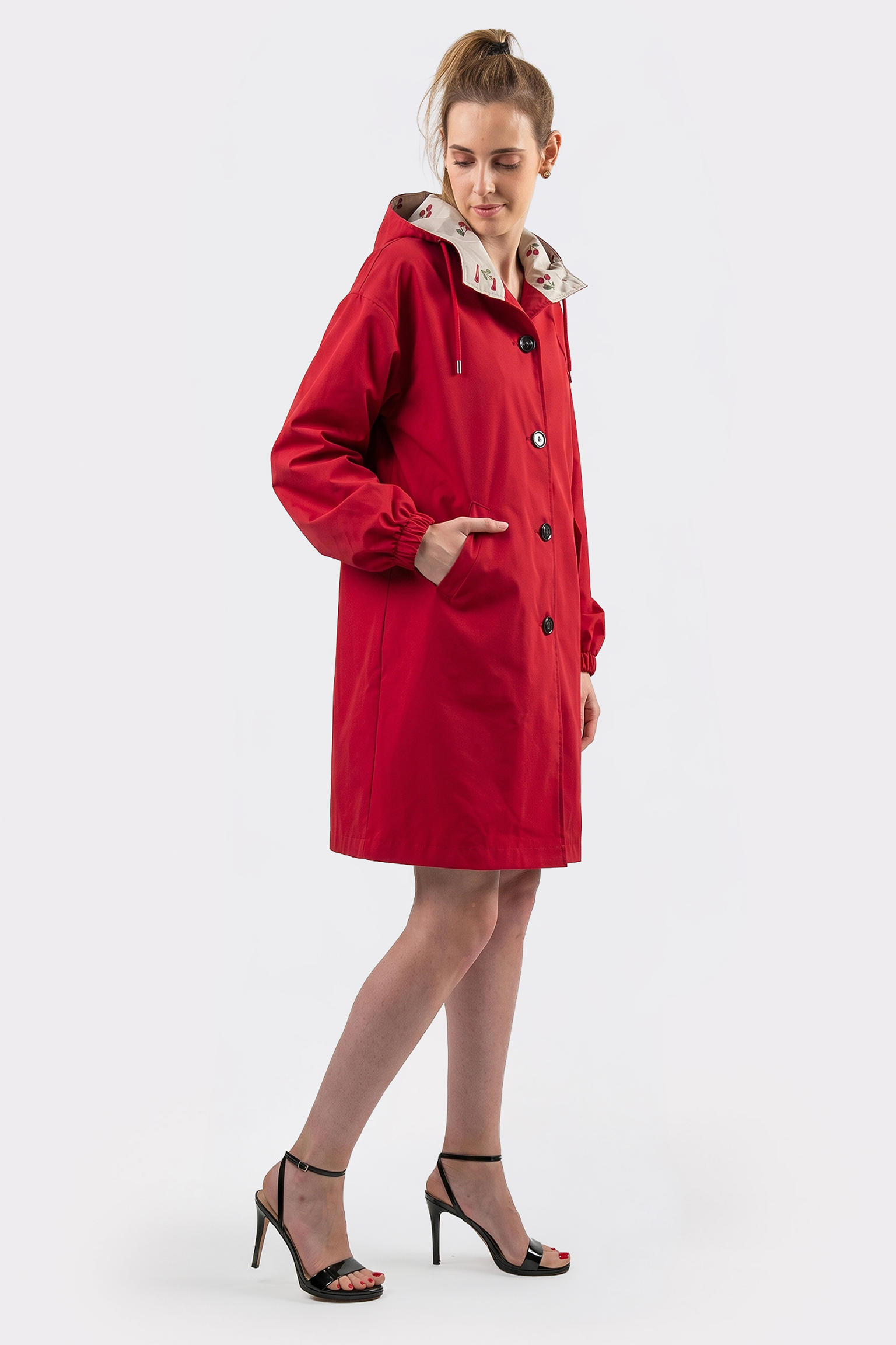 A person stands against a plain light background wearing the Icon Windbreaker Rain Jacket, a knee-length red hooded coat with black buttons and side pockets, paired with black high-heeled sandals.