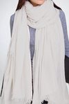 A person is wearing a large, cream-colored Color-POP Scarf by looselyboho draped around their neck and shoulders. The all-weather scarf has a soft, woven texture with frayed edges. The individual is also wearing a gray sweater. The background is plain and light-colored.