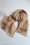 A soft beige Color-POP Scarf with frayed edges from looselyboho is laid out on a flat white surface. The fabric appears light and slightly sheer, with a loose and draped arrangement. The scarf has an elegant, casual look suitable for various outfits, making it an ideal all-weather accessory to pair with Wjackets.