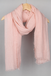 A soft, light pink looselyboho Color-POP Scarf is elegantly draped over a wooden hanger. The scarf has a slightly textured, crinkled appearance with frayed edges, giving it a cozy and casual look. The neutral background emphasizes the gentle and delicate color of this all-weather accessory.