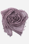 A lavender-colored, Lightweight Linen Scarf from looselyboho is arranged in a swirled, rose-like pattern against a plain white background. The scarf has a delicate, slightly frayed texture that adds an artisanal charm, creating a soft and airy appearance.