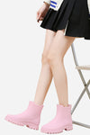 A person sits on a chair, wearing a black skirt and light pink looselyboho Non-Slip Rain Boots. Their hands are resting on their thighs, and their face is not visible. The background is plain white.