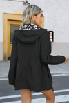 A person with blonde hair is walking across a street in a FLORALSHIFT COAT: THE REVERSIBLE RAINCOAT by looselyboho. They are facing away from the camera. The background features a white brick building with a wooden door and a partially visible sign that reads "Viva.