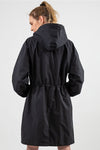 A person with a ponytail is seen from behind wearing The WJacket's Water Resistant Oversized Hooded Windbreaker Rain Jacket, which embodies timeless style. The raincoat features an adjustable waist with a drawstring, slightly cinching it for a versatile fit. The background is plain and light-colored.