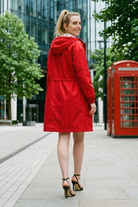 Wearing the Icon Windbreaker Rain Jacket, a person stands on a city sidewalk, looking back. The background features trees, modern glass buildings, and a red British phone booth.