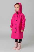 A young child in the looselyboho Bright Berry Kids’ Windbreaker with black buttons and pockets, paired with light pink leopard print rain boots, smiles against a plain light gray background.