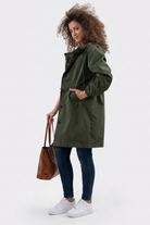 A person stands against a plain background wearing the Icon Windbreaker Rain Jacket in olive green, blue jeans, white sneakers, and holding a brown leather tote bag in their left hand.