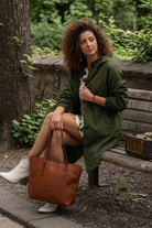 A woman with curly hair sits on a wooden bench in a park, wearing the Icon Windbreaker Rain Jacket, a patterned dress, and white boots. She holds a brown leather tote bag and looks to the side, with a vintage radio beside her.