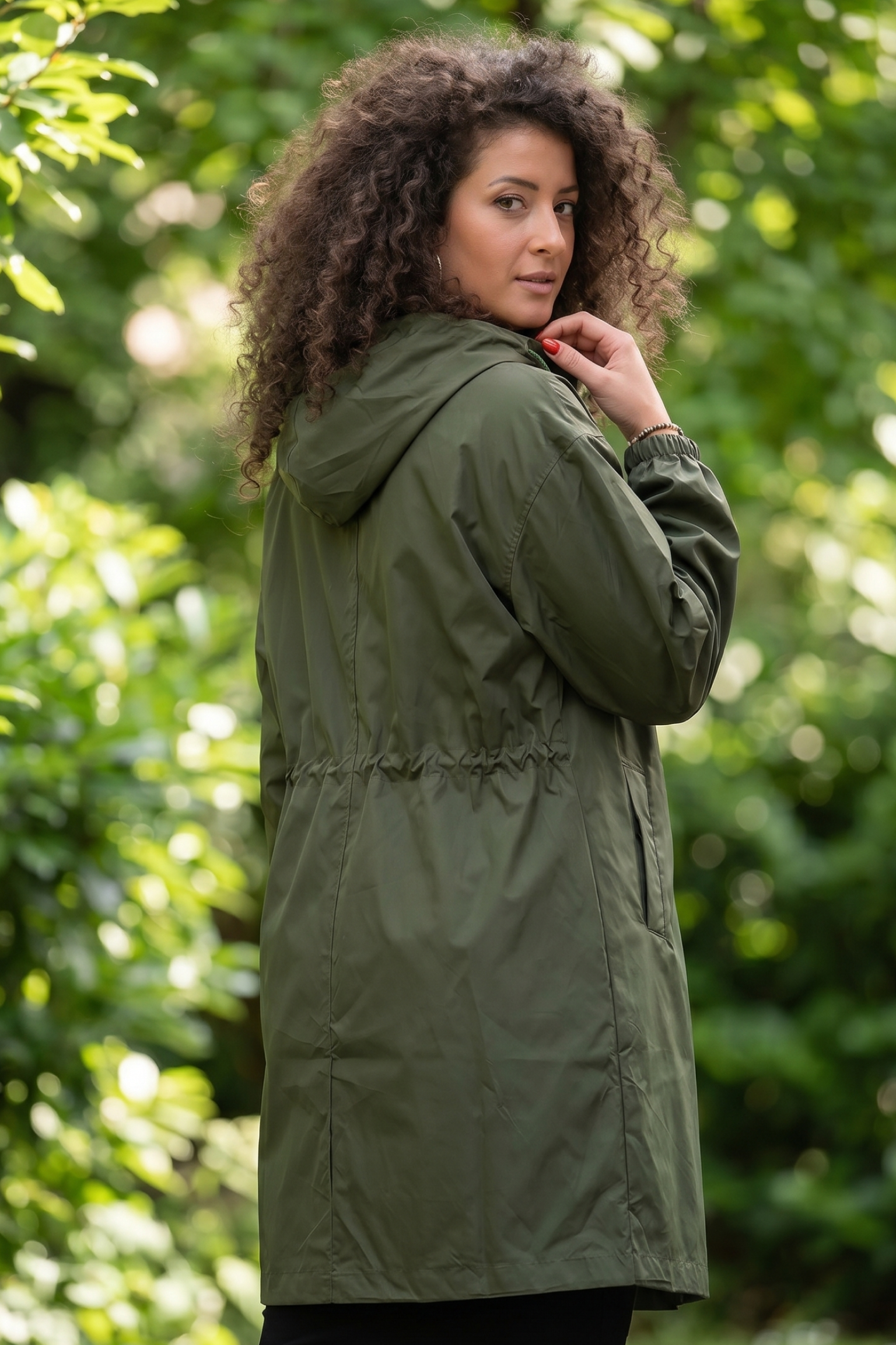 A woman with curly hair stands outdoors, looking over her shoulder while wearing the Icon Windbreaker Rain Jacket in olive green. Lush green foliage fills the background.