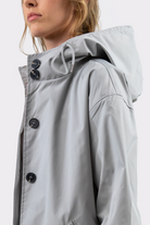 Someone in the Icon Windbreaker Rain Jacket—a light gray, matte-finish hooded raincoat with black buttons—shown from the shoulders up and facing sideways. The hood is up but not covering the head.