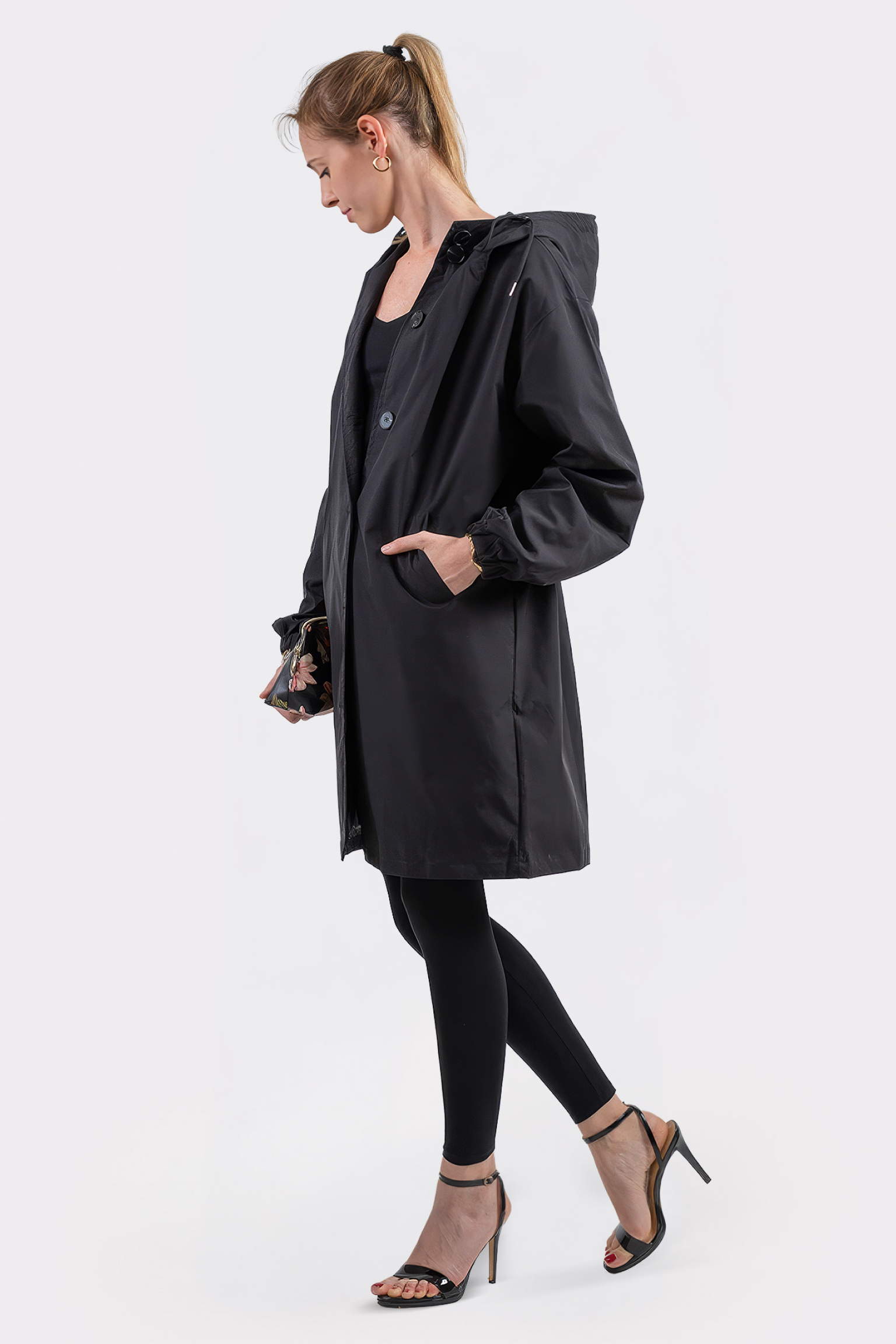 A woman in profile wears the Icon Windbreaker Rain Jacket with black leggings and heeled sandals. Her hair is styled in a ponytail, one hand in her pocket, standing against a plain white background.