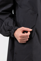 A close-up shows a person wearing the Icon Windbreaker Rain Jacket in black, with their right hand holding the edge of its slanted pocket. The jacket features an elastic cuff at the wrist, set against a plain light background.