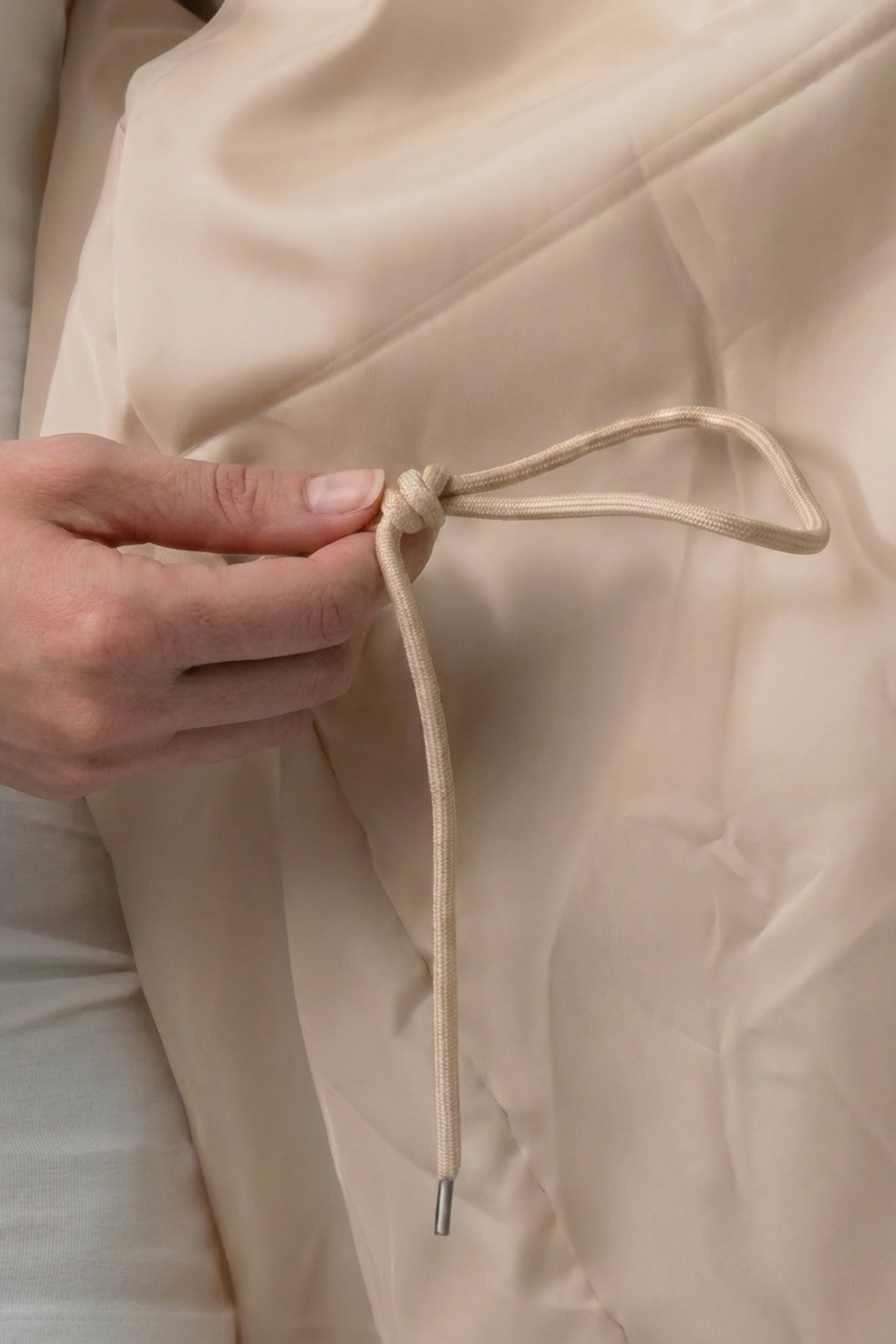A hand holds a beige drawstring tied in a bow against the fabric of the Icon Windbreaker Rain Jacket.