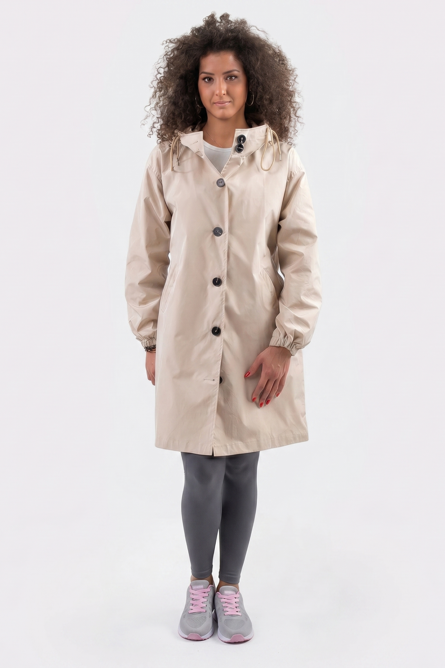 A person with curly hair stands facing forward, wearing the beige Icon Windbreaker Rain Jacket, gray leggings, and gray sneakers with pink laces against a plain white background.