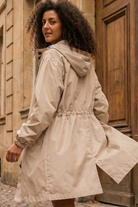 A person with curly hair wearing the Icon Windbreaker Rain Jacket in light beige stands outdoors in front of a wooden door and stone building, looking back over their shoulder.