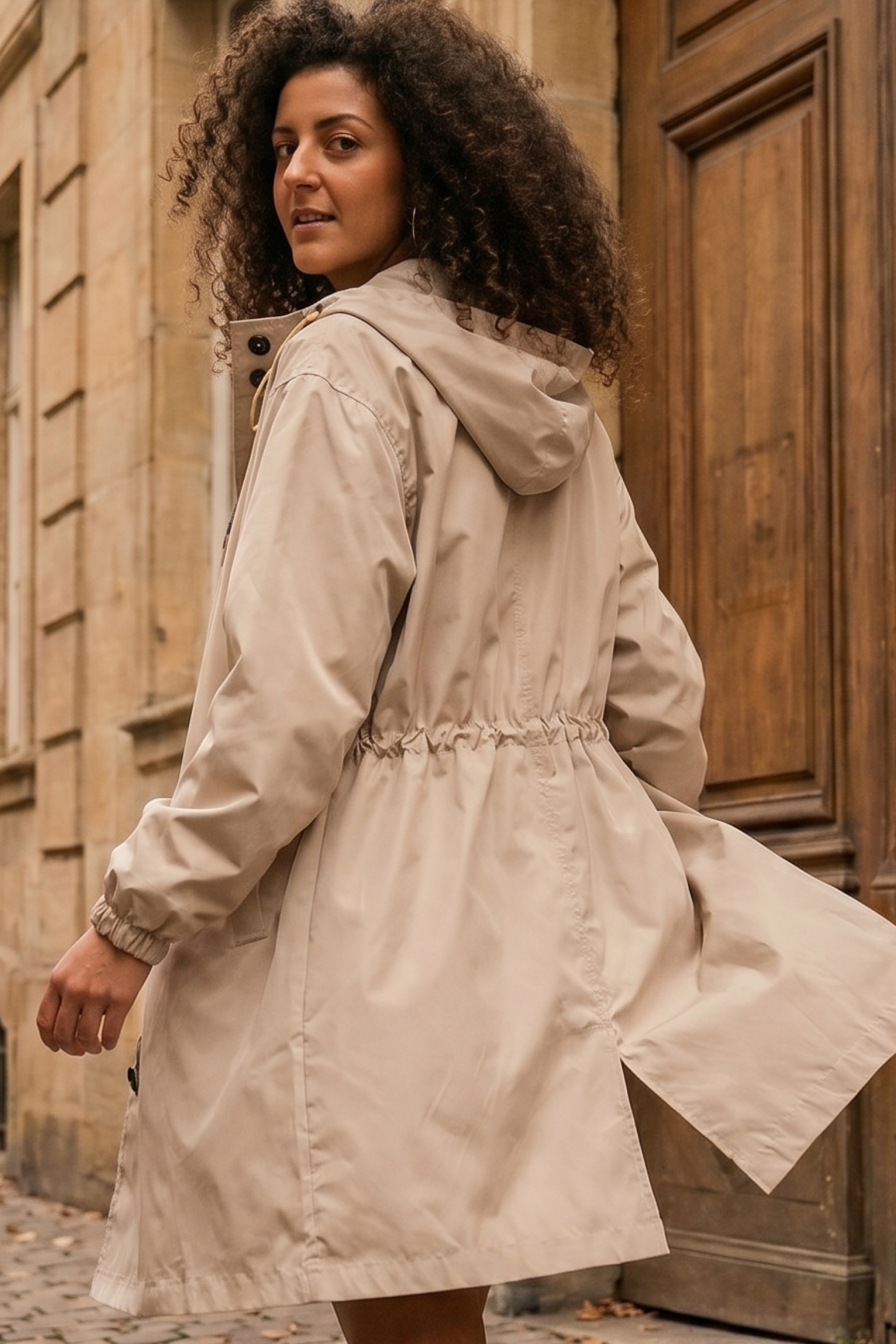 A person with curly hair wearing the Icon Windbreaker Rain Jacket in light beige stands outdoors in front of a wooden door and stone building, looking back over their shoulder.