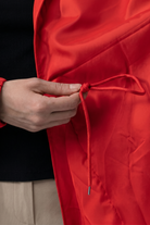 A person ties the drawstring inside the bright red Icon Windbreaker Rain Jacket while wearing beige pants and a black shirt. The photo highlights their hands and the jacket's interior.