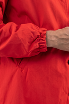 A close-up of a person wearing the Icon Windbreaker Rain Jacket, featuring a bright red color and elastic cuff. Their arm is bent, showing only part of their hand and forearm against the jacket.