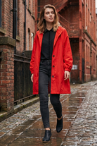 A woman wearing the Icon Windbreaker Rain Jacket, a black shirt, black jeans, and black shoes walks on a wet cobblestone street between red brick buildings.
