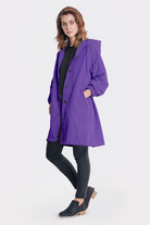 A woman faces forward against a plain background, wearing the Icon Windbreaker Rain Jacket in purple with a hood, paired with a black top, black skinny jeans, and black low-heeled slip-on shoes. One hand is casually in her pocket.