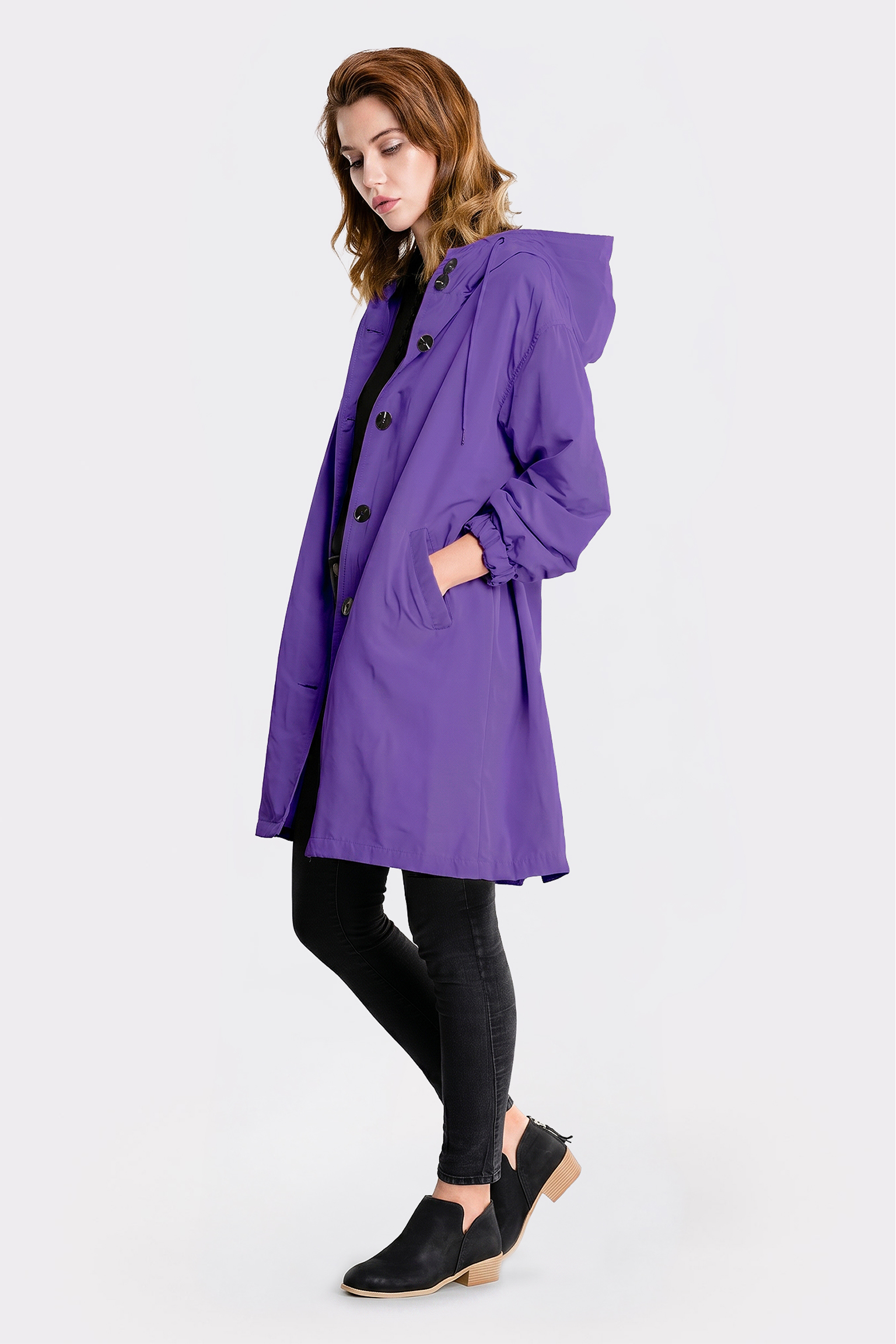 A person stands sideways wearing the knee-length Icon Windbreaker Rain Jacket in purple with large buttons and pockets, paired with black pants and low-heeled black slip-on shoes. The background is plain white.