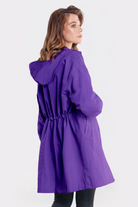 A woman with light brown hair wears the Icon Windbreaker Rain Jacket, a long purple hooded coat. She stands facing slightly away from the camera with her left hand in her pocket against a plain white background.