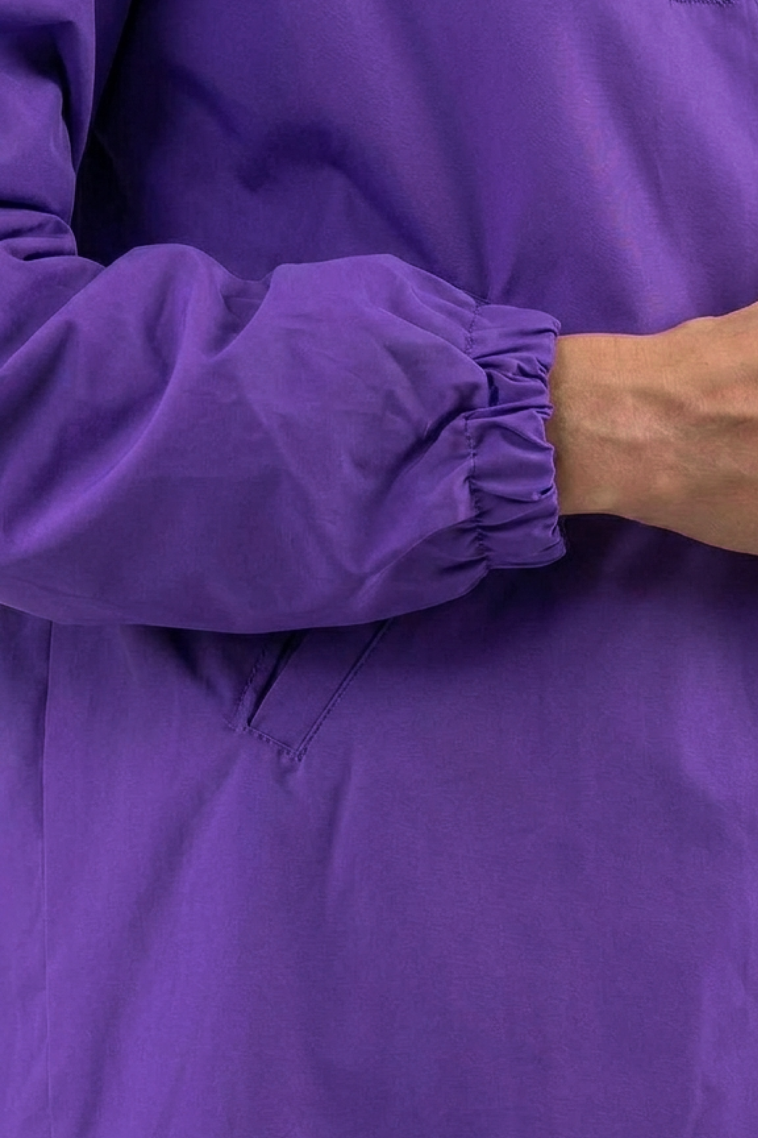 A person is shown from shoulder to waist wearing the Icon Windbreaker Rain Jacket in purple, featuring elastic cuffs, a side pocket seam, and one hand partially visible.