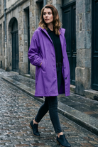 A woman in the Icon Windbreaker Rain Jacket, a black top, dark jeans, and black shoes walks on a wet cobblestone street beside stone buildings with dark doors.