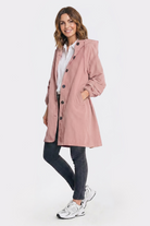 A woman with shoulder-length brown hair smiles as she faces forward, wearing the Icon Windbreaker Rain Jacket in light pink over a white shirt, paired with dark pants and white sneakers. One hand is in her jacket pocket. The background is plain white.