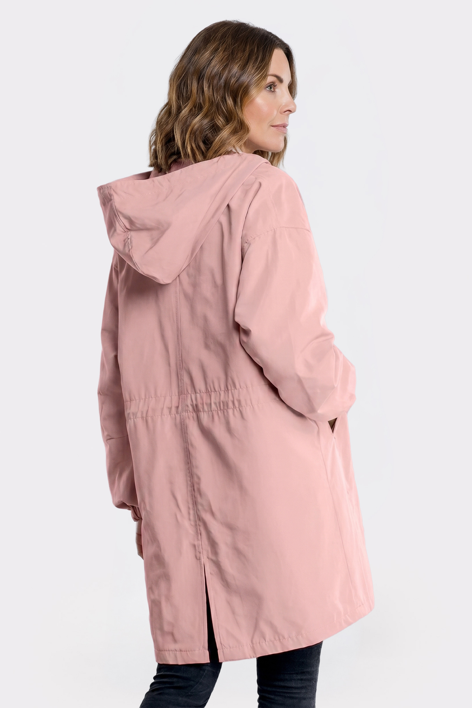 A woman with wavy brown hair is seen from the side and back, wearing the Icon Windbreaker Rain Jacket in light pink, mid-thigh length, standing against a plain light background with her hands relaxed by her sides.