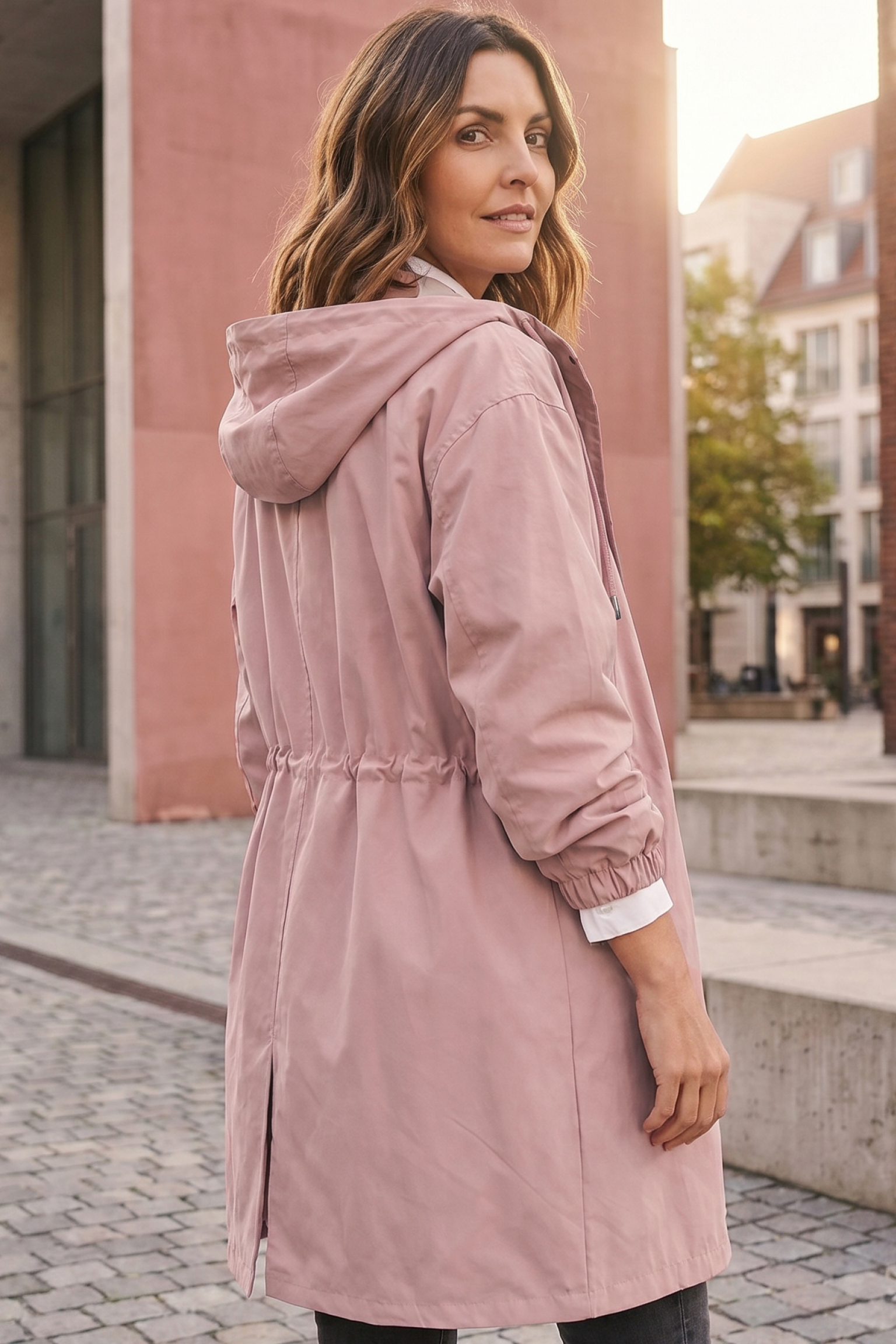 A woman with wavy brown hair stands on a cobblestone street, wearing the Icon Windbreaker Rain Jacket in light pink and looking over her shoulder. Buildings and trees appear in the background.