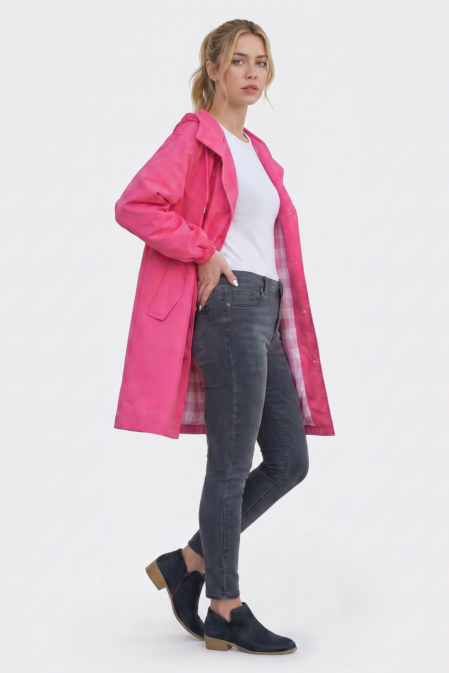 A woman faces slightly to the side in an Icon Windbreaker Rain Jacket—bright pink with a plaid lining—worn open over a white t-shirt, gray skinny jeans, and navy ankle boots. The background is plain white.