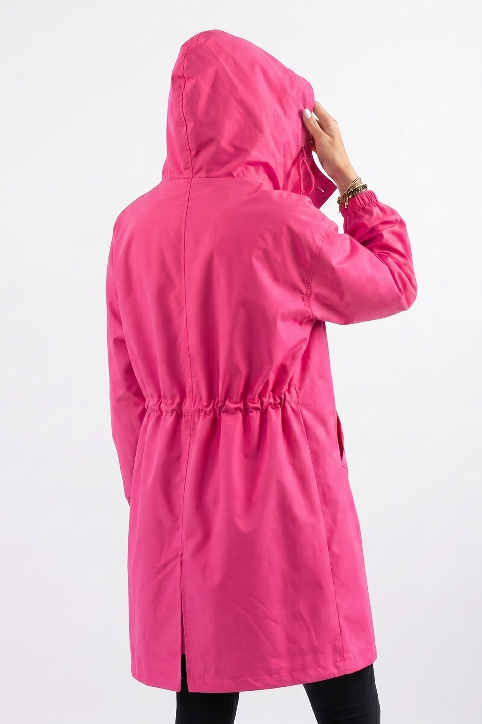 A person is seen from the back touching the hood of an Icon Windbreaker Rain Jacket, featuring a bright pink color and an elastic waistband, against a plain white background.