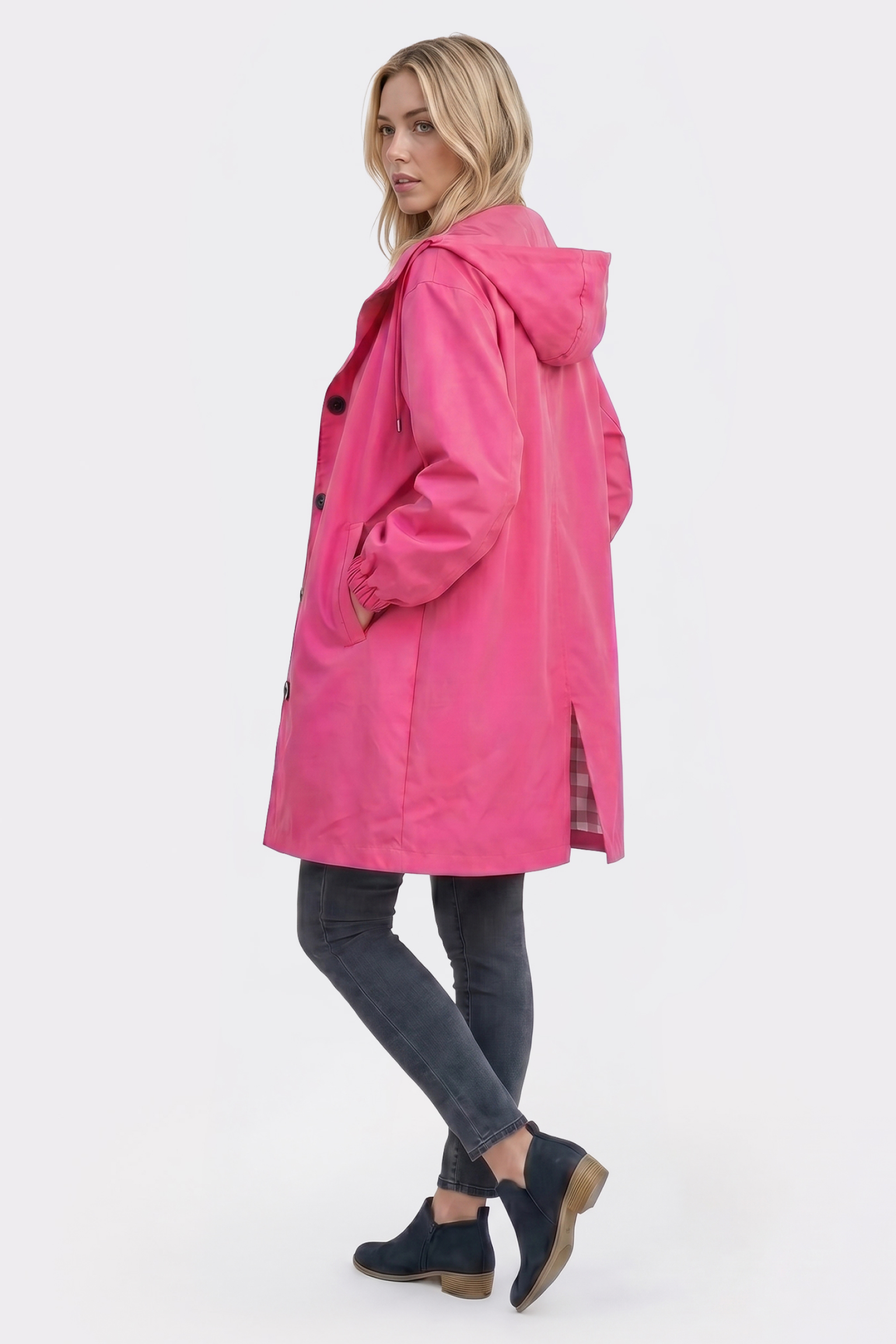 A woman stands slightly turned away, wearing the Icon Windbreaker Rain Jacket in bright pink with dark jeans and black ankle boots. Her left hand is in her pocket as she looks over her shoulder against a plain white background.