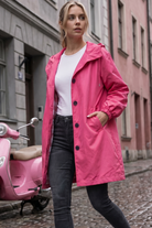 A woman in an Icon Windbreaker Rain Jacket, white shirt, and dark jeans walks down a cobblestone street. A pink scooter is parked nearby, with tall-windowed buildings lining the street.