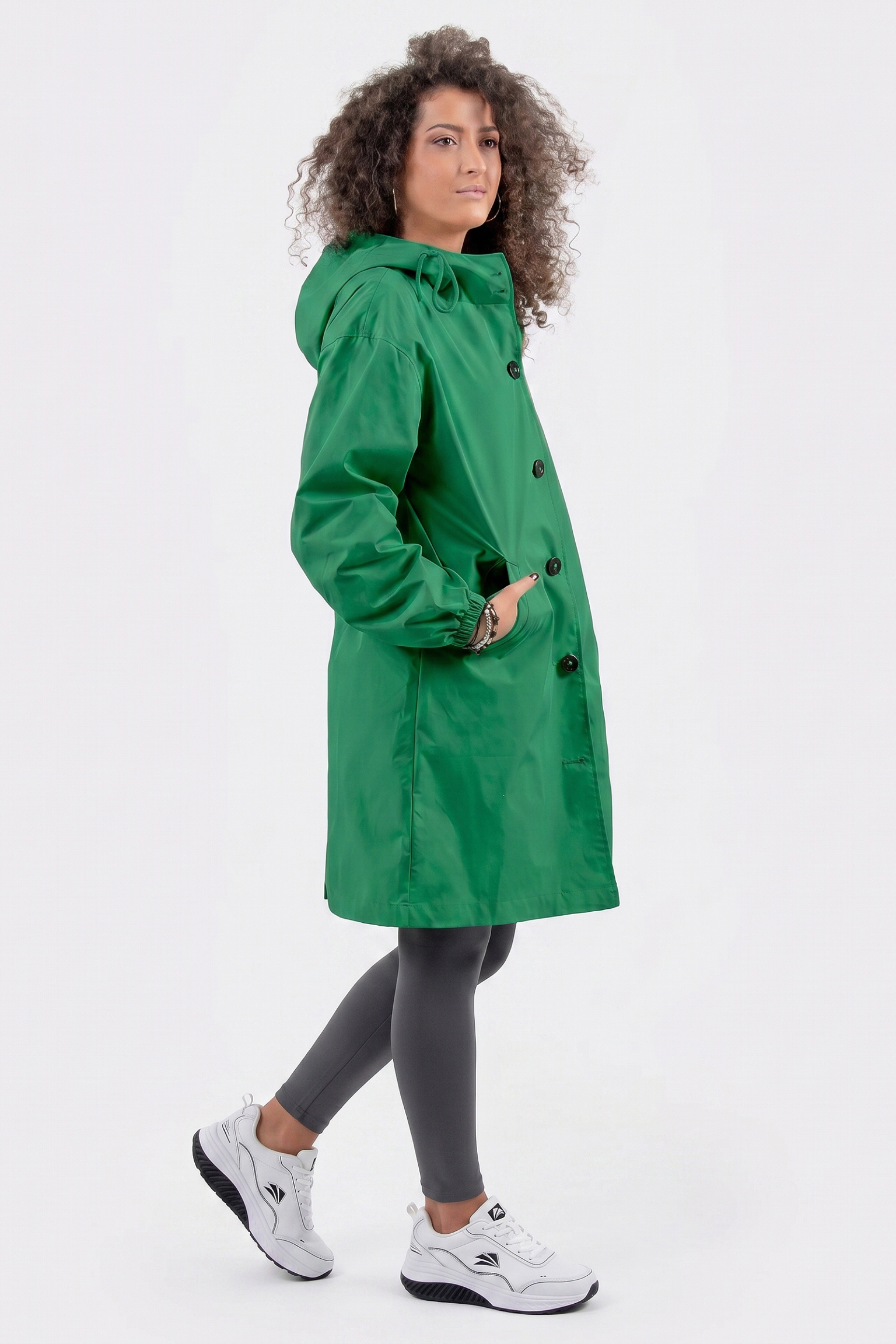 A person with curly hair is wearing the Icon Windbreaker Rain Jacket in green, paired with gray leggings and white sneakers. They stand against a plain white background, facing slightly to the side.