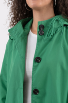 A person wears the Icon Windbreaker Rain Jacket in green with black buttons over a white ribbed shirt. The upper body is shown, face partially visible, with curly hair and hoop earrings.